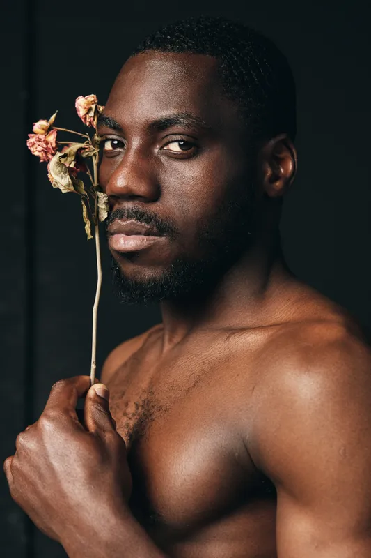 African man with dark skin holding dried flower close to face in creative conceptual visual style. Portrait, bare chest and serious expression on black background for artistic photography.