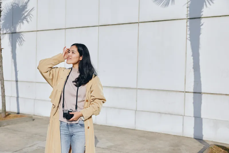 Woman with camera shields eyes while standing outdoors beside a white wall, wearing a beige coat and denim, capturing street photography in bright sunlight and soft palm shadows.