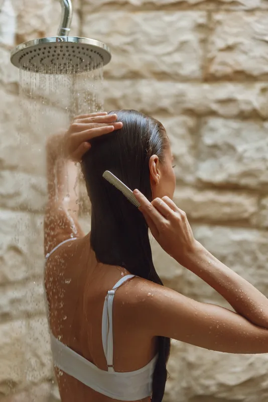 Woman combing wet hair under shower water with natural stone wall background, calm and refreshing morning routine emphasizing self-care and hygiene.