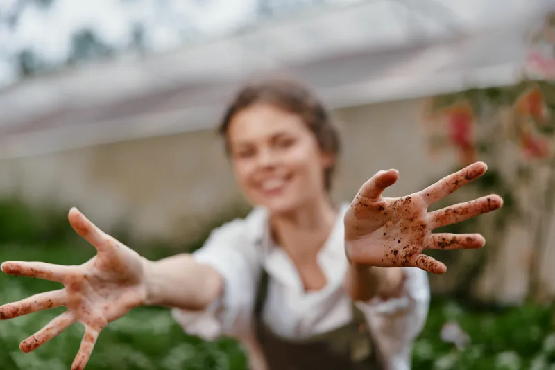 Young woman with dirt covered hands reaching out, smiling, in a lush garden setting, symbolizing gardening, nature, and hands on experience