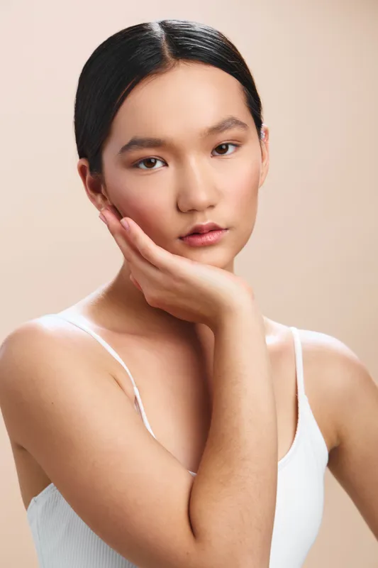 Asian woman showcases a calm, thoughtful pose in a clean studio setting, her hand resting on her cheek, conveying poise, focus, and quiet confidence suitable for versatile portrait use.