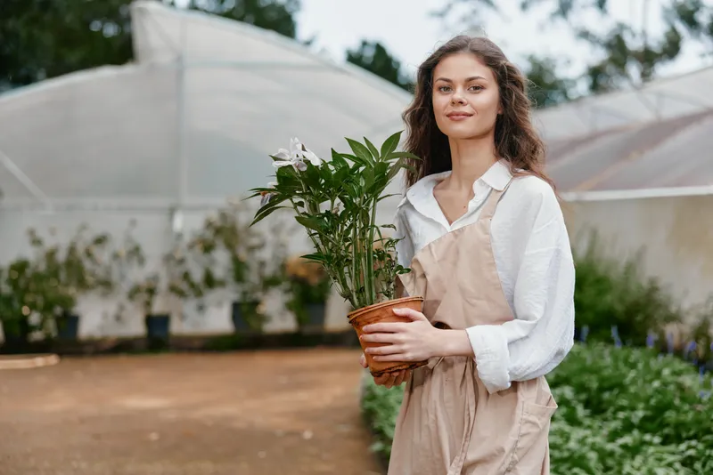 Young woman in a garden greenhouse, holding a potted plant, dressed in beige apron and white shirt, joyful expression in a natural setting