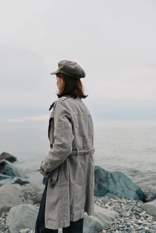 Woman in grey coat and hat stands on rocky beach facing calm sea under cloudy sky. Concept for solitude, contemplation, and autumn fashion outdoors.