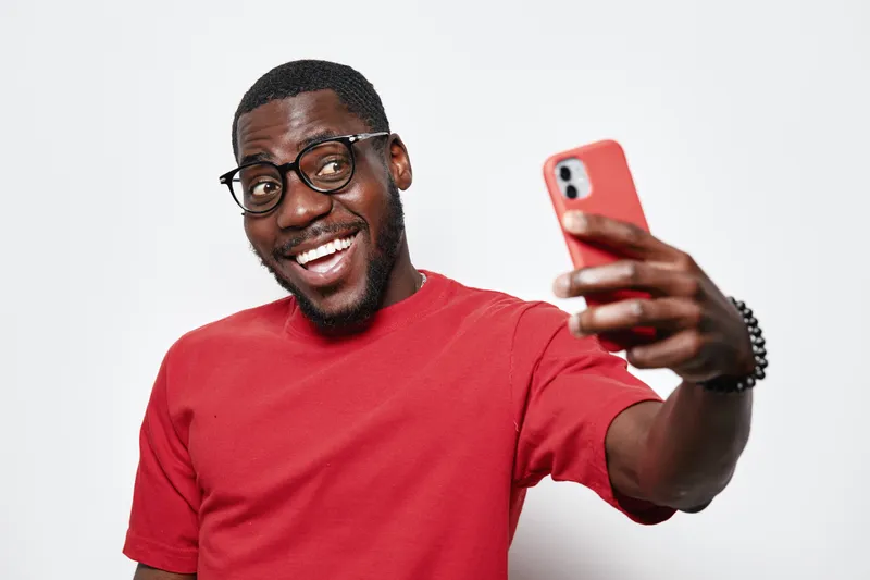 African American man with dark skin and glasses wearing red shirt smiling and taking selfie with smartphone on white background. Happy young male holding phone for photo.