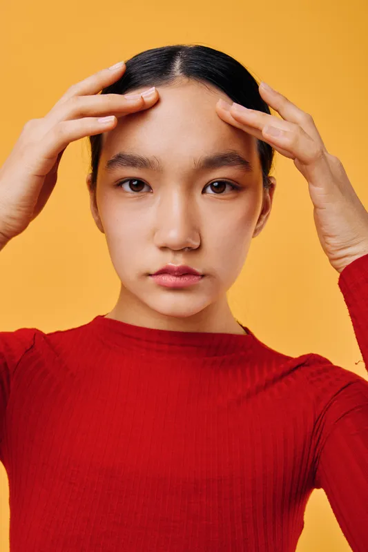 Young woman with clear skin and black hair in red sweater touching forehead with fingers on yellow background for skincare, beauty, health and wellness concept portrait.