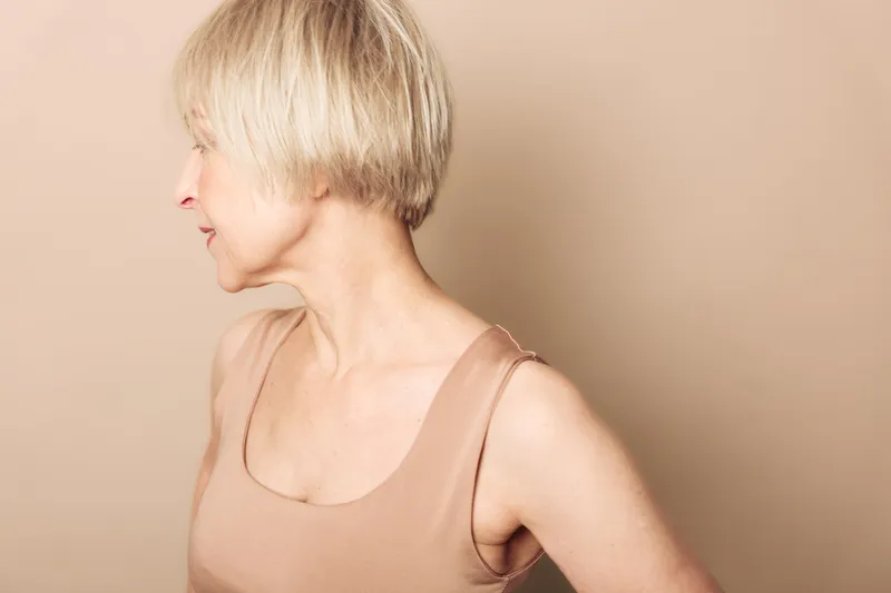 Profile of a mature woman with short blonde hair wearing a beige tank top against a neutral background. Concept of aging, natural beauty, and simplicity in portrait photography.