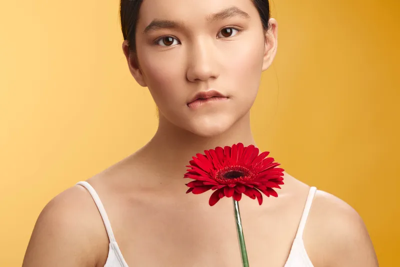 Asian woman holds a red flower near her chest, facing the camera with a calm expression. The warm yellow background and soft lighting create a serene, contemporary portrait mood.