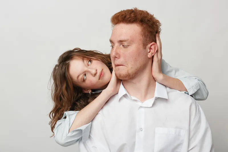 Young couple in white shirts poses against light background. Woman embraces man from behind, looking at him with affection. Concept of love, relationship, intimacy, and connection.