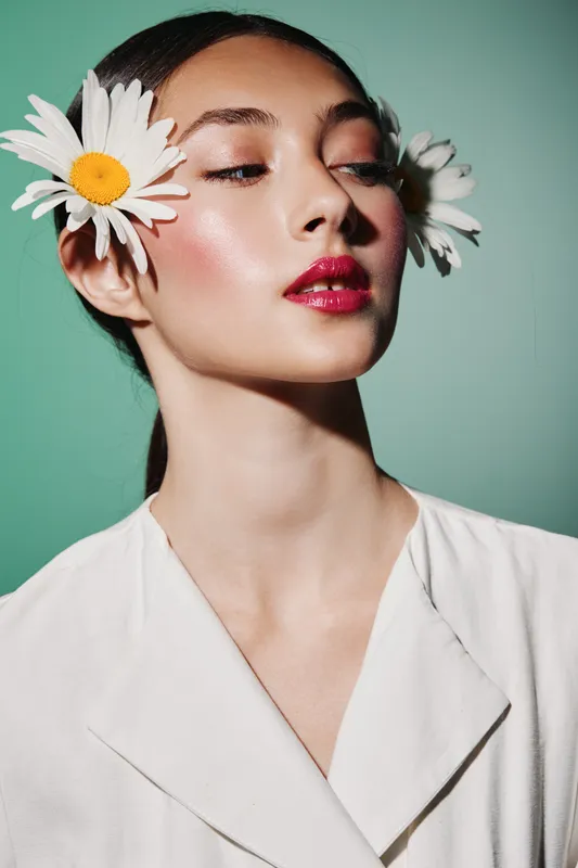 Beauty portrait of a stylish woman with glossy red lips and radiant skin adorned with white daisies on a pastel green background. Concept of fashion and creative beauty.