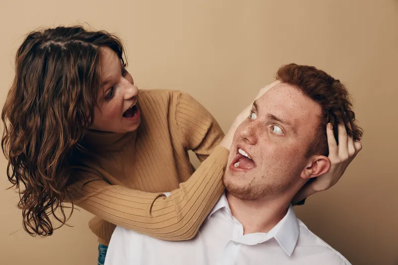 Surprised man with red hair and freckles reacts to playful woman grabbing his head. Casual indoor scene with beige background, showing fun interaction and friendly relationship between adults.