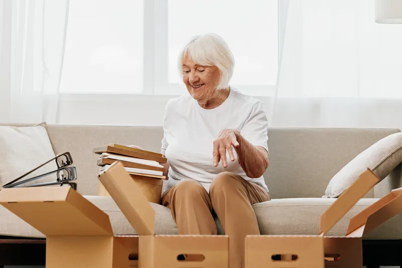 Пожилая женщина сидит на диване дома среди коробок