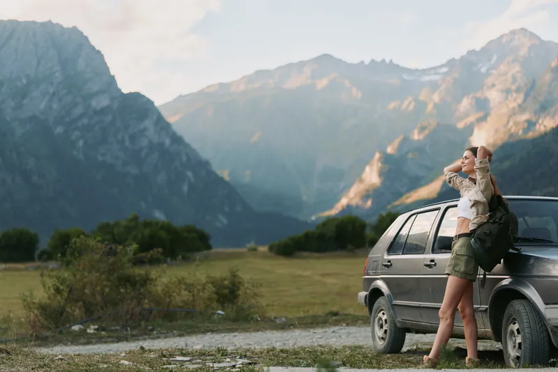 Young woman with a backpack stands beside a car in a rugged mountain landscape, gazing into the distance with a sense of adventure, freedom, and anticipation as dramatic peaks loom behind her.