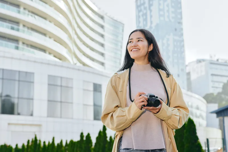 Camera in hand, a smiling woman stands in an urban outdoor setting with modern buildings behind. Casual coat and sweater under soft daylight convey travel, photography, and city exploration for stock