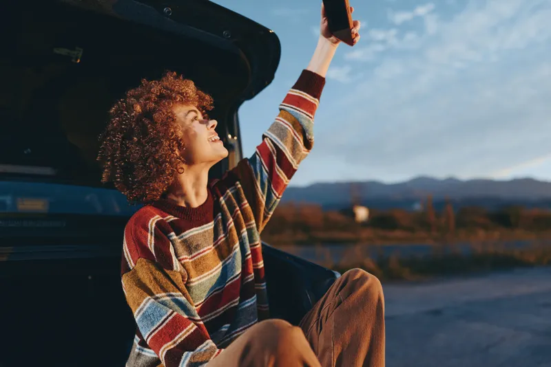 Woman lifestyle travel, curly woman by car open trunk, outdoors lifting hand, striped sweater and brown pants, sunset light, mountains in distance, calm roadside moment