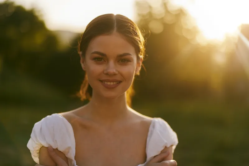 Young woman in a white dress smiling warmly during golden hour, embodying joy and serenity in a vibrant outdoor setting