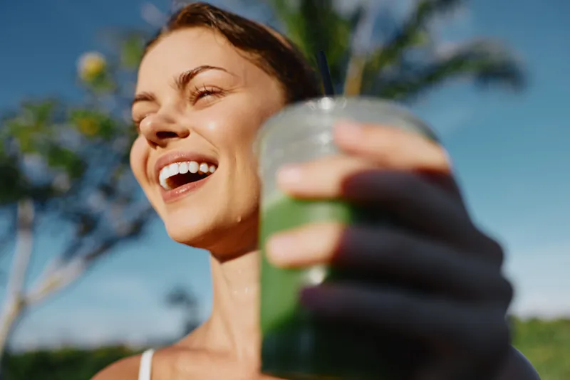 Smiling woman outdoors holding a green smoothie, sunny day, natural light, wellness lifestyle, healthy eating, self care moment, closeup feel, joyful mood, vibrant colors.