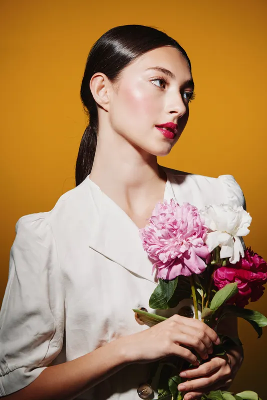 Elegant young woman with glossy makeup and vibrant lipstick poses with pink and white flowers on a bold yellow background. Concept of beauty creative fashion glamour.