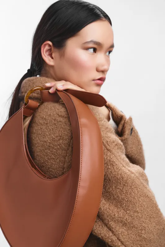 asian woman wearing a cozy coat with a brown leather bag over her shoulder, posed against a neutral backdrop to highlight fashion, confidence, and contemporary style in a studio portrait.