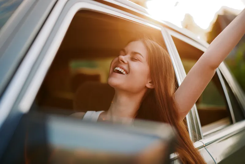 Joyful young woman with long hair enjoying a summer road trip by the window of a car, celebrating freedom and adventure in nature.