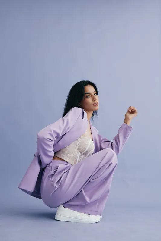 young woman in lilac suit crouching confidently against plain purple background wearing white sneakers and lace top, stylish casual outfit with modern urban vibe