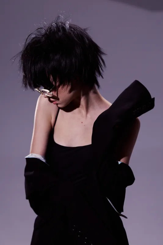 Young woman with messy hair wearing glasses and a black outfit posing against gray background.