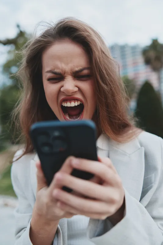 Frustrated woman yelling at smartphone outdoors expressing emotional stress with blurred city background and casual jacket in autumn tone