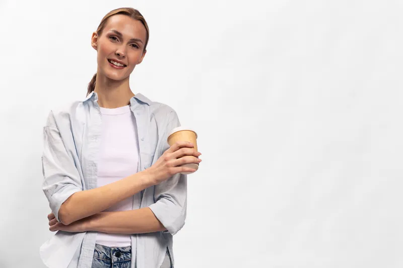 Smiling young woman in casual clothes holds a takeaway coffee cup, standing against a plain light background. Concept of coffee break, relaxation, and casual lifestyle.