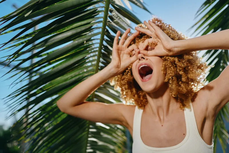 young woman with curly hair posing joyfully near palm leaves, wearing white tank top and expressing summer happiness outdoors under bright sunlight