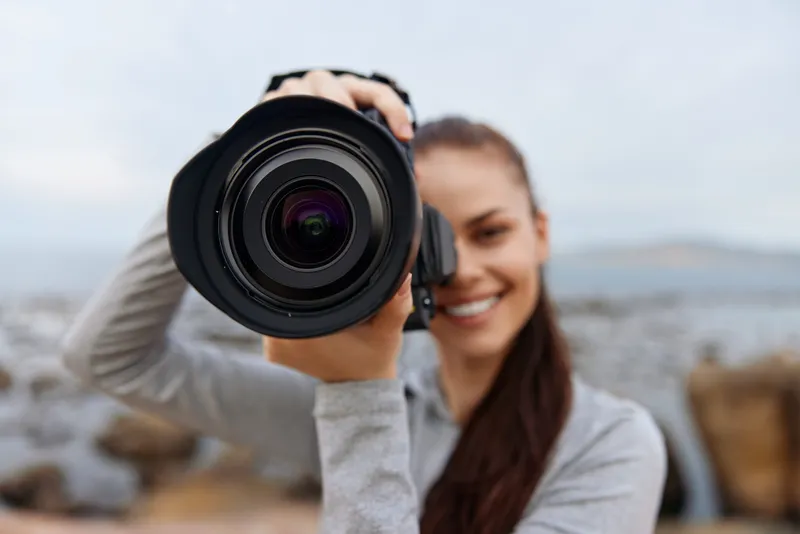 Young woman photographer smiling while holding a camera on a rocky beach, showcasing creativity, passion, and confidence