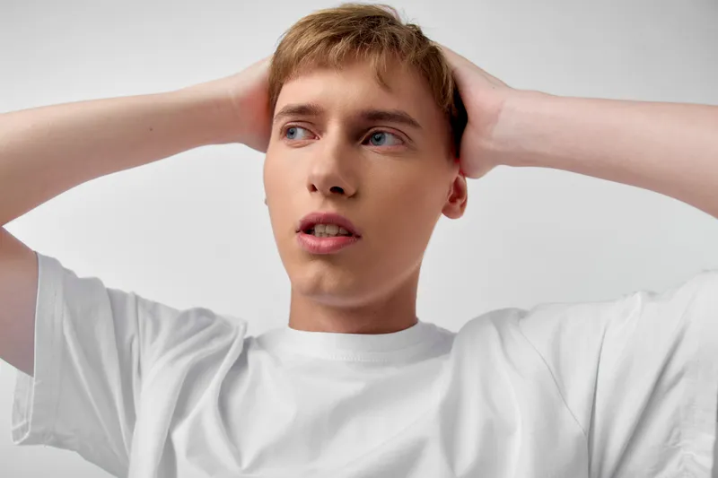 Young man with hands on head, white shirt, surprised expression in a clean studio, conveying tension, confusion and contemplation in a minimal, contemporary setup