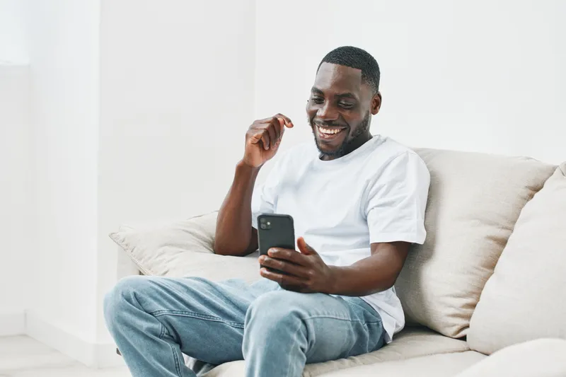 Happy Black man sitting on a couch using a smartphone with a joyful expression, dressed in casual white t shirt and blue jeans, in a bright interior setting
