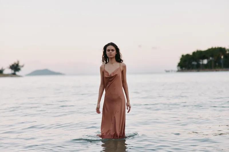 A model stands in shallow sea water wearing a silky brown dress, calm expression, soft lighting, gentle waves, distant horizon, creating a serene coastal portrait.