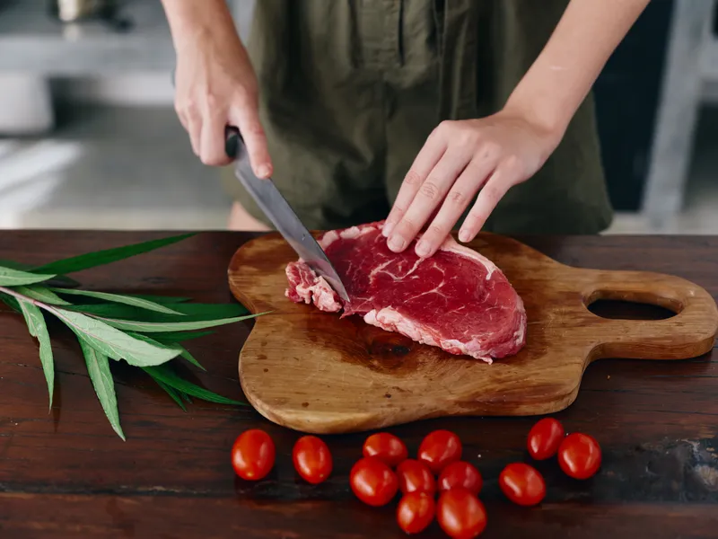 Женщина с ножом в руке нарезает свежее стейковое мясо для жарки на кухне