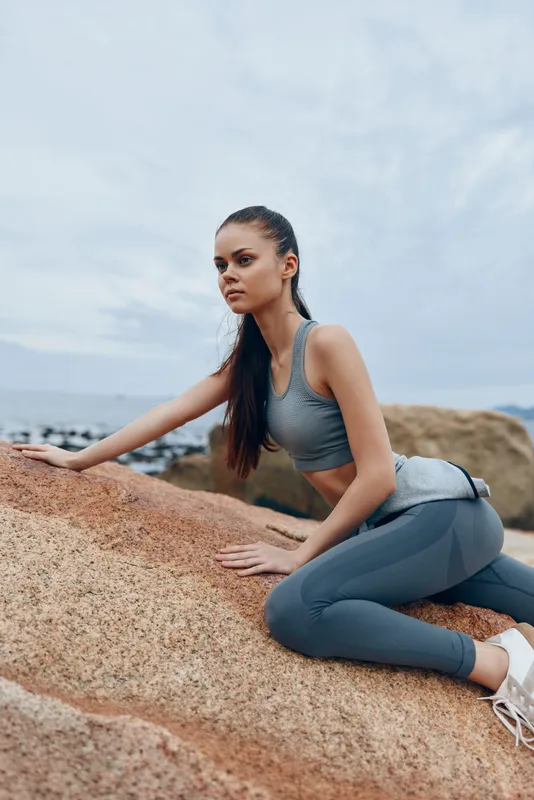 Young woman in athletic wear posing on rocky beach, showcasing fitness lifestyle, confidence, and natural beauty against a serene coastal background
