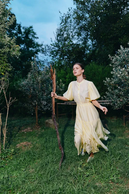 A woman in a flowing yellow dress walks through a grassy field holding a wooden staff, surrounded by trees for a serene outdoor scene at dusk.