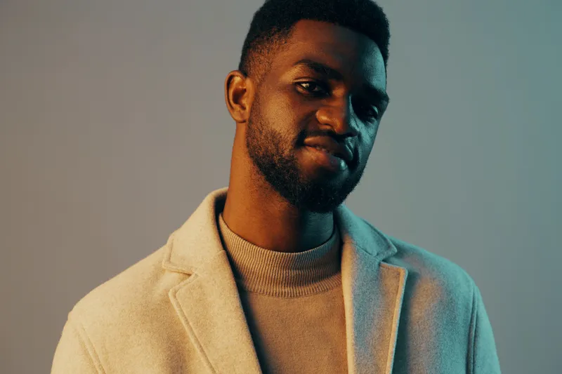 Stylish young man with a confident expression, wearing a light coat against a contrasting blue and beige background, showcasing contemporary fashion and personal style