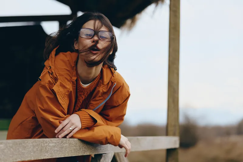 Woman lifestyle outdoors in an orange jacket and glasses wearing a mask, leaning on a wooden bench, warm sunlight, calm smile, soft breeze, blurred rural background, cool season mood