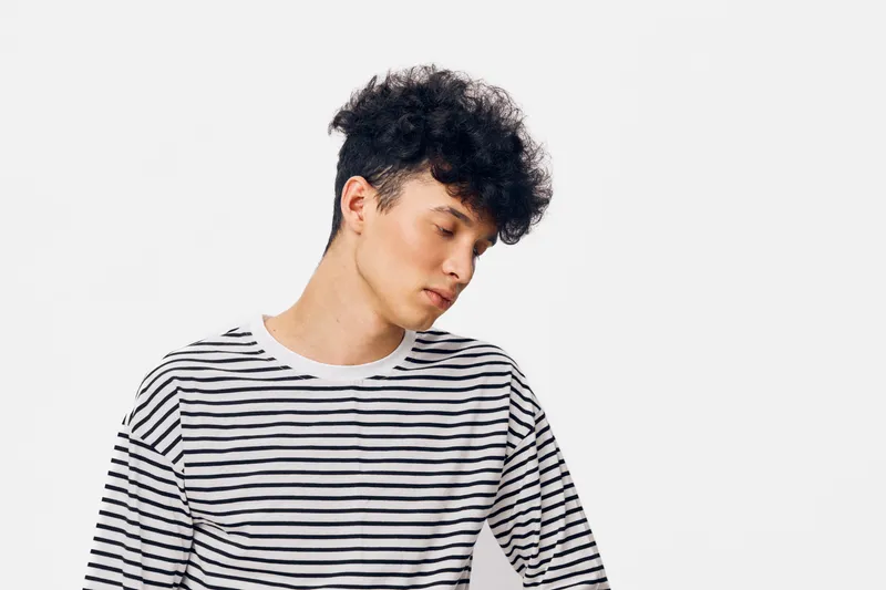 young man in striped shirt looking down pensively with curly hair on white background, casual pose and natural expression