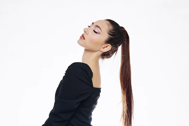 Young woman with long ponytail and bold purple eye makeup poses confidently in off-shoulder black outfit. Clean white background highlights beauty fashion and creative style.
