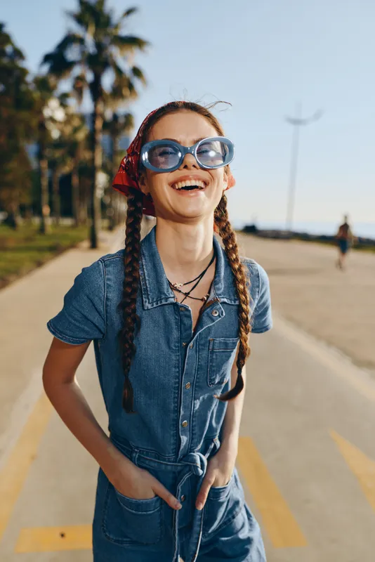 smiling woman wearing blue sunglasses and denim jumpsuit standing outdoors on sunny day with palm trees and clear sky background