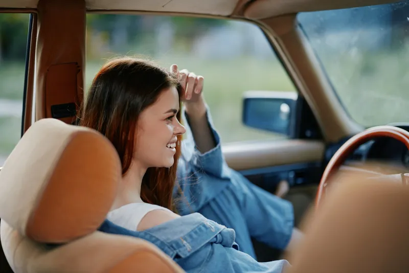 Young woman smiling in a vintage car, enjoying a sunny day in nature, wearing casual jeans and a denim jacket, representing freedom and joy.