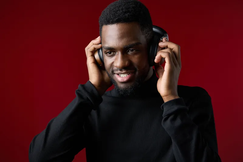 Happy young Black man enjoying music with headphones in a stylish black outfit on a vibrant red background