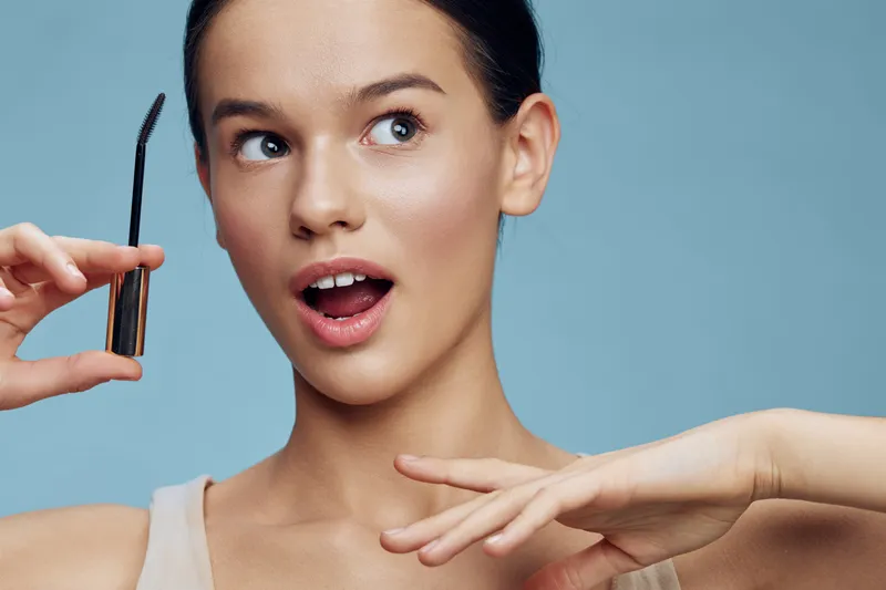 Excited young woman applying mascara, showcasing makeup routine with a surprised expression against a light blue background, emphasizing beauty and self care
