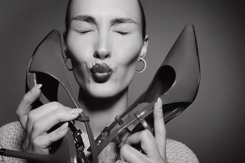 Close-up black and white portrait of a young white woman with closed eyes holding high heel shoes near her face, expressing affection and fashion concept in studio light.