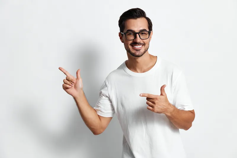 Full body smiling cheerful young man wearing white t-shirt and glasses, gesturing with hands while looking at the camera against a plain white backdrop.