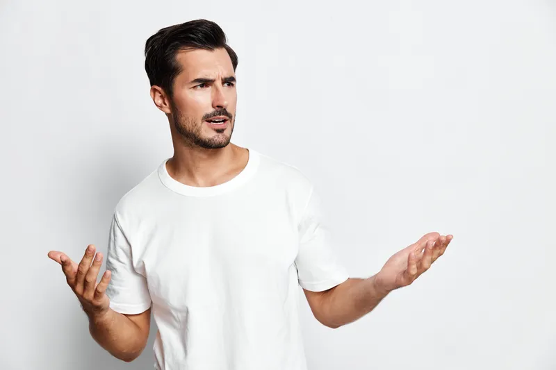 Confused man with stylish short hair wearing a plain white t-shirt, looking at the camera with a questioning expression, isolated on a soft gray background.