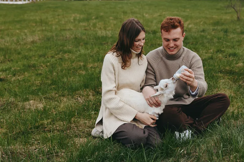 A couple in cozy sweaters sits on grass outdoors, gently cradling a small goat kid. Warm light and a serene field backdrop evoke lifestyle, family bond, and care for animals, together in nature.
