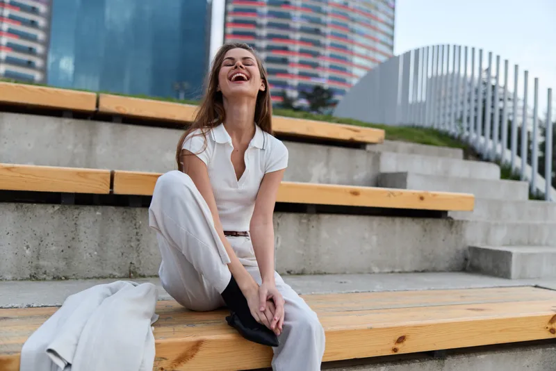 Young woman in casual white outfit laughing joyfully sitting on urban wooden steps with modern city background lifestyle fashion emotion woman casual outdoor