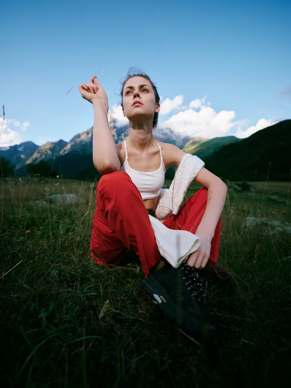 Outdoor landscape with a confident woman crouching in a green field, red pants and white top, fist raised, blue sky and distant mountains, conveying resilience and freedom