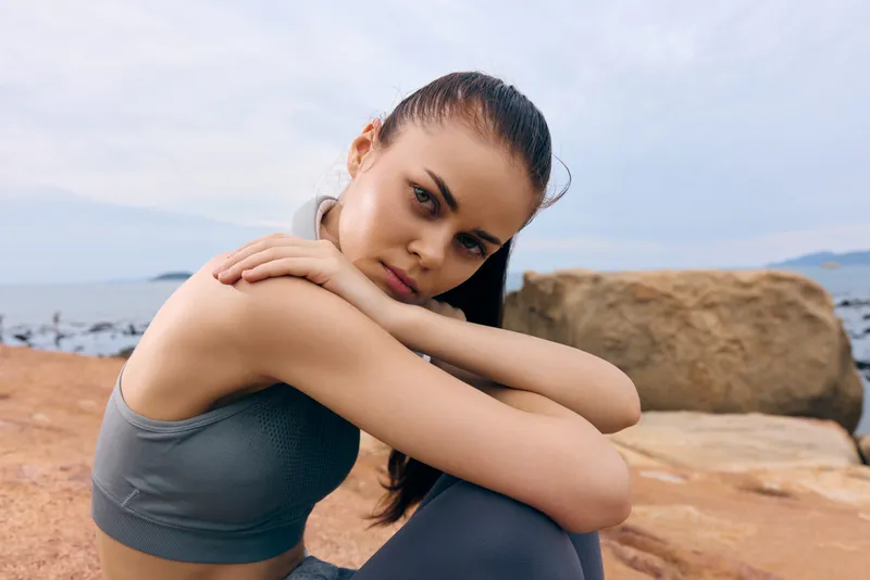 Young woman in sporty attire, sitting on rocks by the sea, expressing determination and confidence, with a soft color palette and a serene background